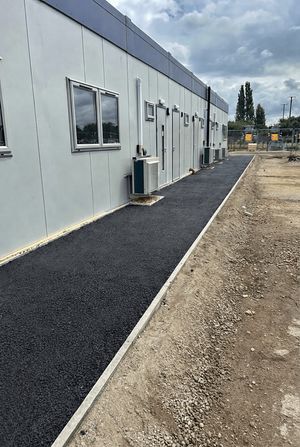 A newly constructed modular office building with a fresh asphalt footway and external air conditioning units, adjacent to a gravel and dirt area at a development site