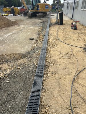 Newly installed drainage channels run alongside a modular office building at a construction site, with heavy machinery and groundwork materials present