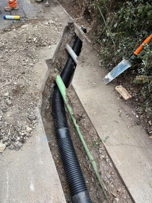 Open trench along a roadside containing black corrugated electrical conduits and a green utility duct, with construction tools nearby, on Anstey Lane, Alton