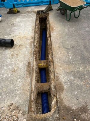Freshly dug trench in a paved surface containing a blue corrugated electrical conduit, with a wheelbarrow of sand and construction barriers, on Anstey Lane, Alton