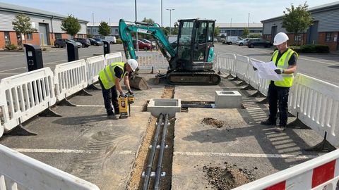 Twin electrical ducts and a draw pit under construction beside a rapid EV charger in an occupied UK commercial car park