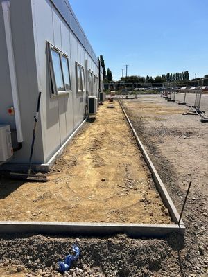 A construction site featuring the side of a modular office building with open windows, bordered by a newly prepared dirt footway and temporary fencing under a clear sky