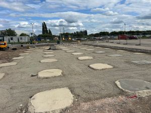 Multiple freshly poured concrete pad foundations arranged in a grid pattern on a construction site, surrounded by gravel, temporary fencing, and construction equipment