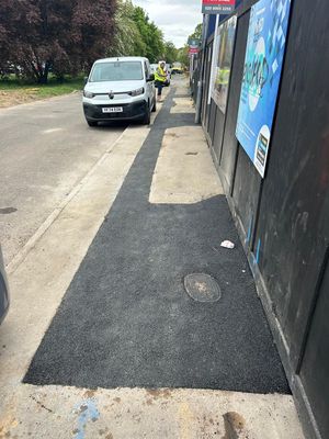 Operative in hi vis inspecting a completed black tarmac footway reinstatement alongside the site hoarding on Aycliffe Road, Borehamwood