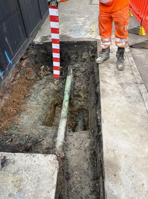 Construction operative in hi vis with a measuring rod beside an open LV cable trench exposing existing services on Aycliffe Road, Borehamwood