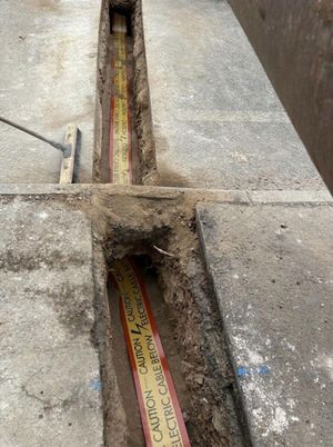 Anstey Lane roadside trench with yellow Caution Electric Cable Below warning tape laid inside, an operative in orange safety gear visible at the top of the frame