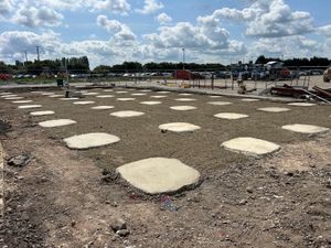 A cleared construction site with several freshly poured concrete pad foundations in a grid, with fencing, vehicles, and construction materials visible under a partly cloudy sky