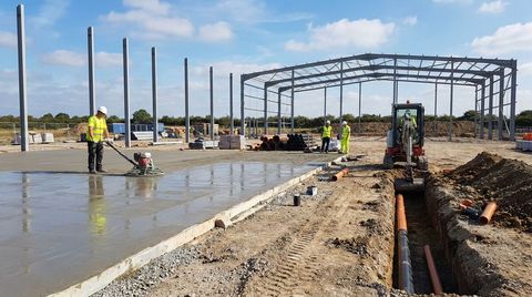 Warehouse groundworks in progress with a fresh concrete floor slab, a drainage trench and steel frame columns going up on a dry day